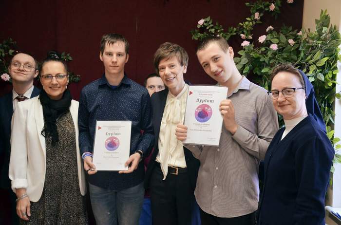 Pokaż zdjęcie: Zdjęcie grupowe sześciu osób stojących obok siebie na tle jasnej ściany. W centrum dwaj mężczyźni prezentują dyplomy z napisem „Dyplom” i kolorową grafiką. Od lewej: kobieta w okularach i jasnej marynarce, młody mężczyzna w granatowej koszuli, w tle twarz kolejnej osoby, starsza kobieta w białej koszuli i ciemnej marynarce, młody mężczyzna w szarej koszuli oraz zakonnica w granatowym habicie. Wszyscy są uśmiechnięci.