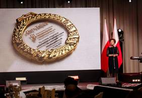Pokaż zdjęcie: Prezez Marlena Maląg zapowiada z mównicy wręczenie nagrody Złotego Tulipana, na ekranie widać tę nagrodę - okrągły szklany medal w złoconej otoczce z grawerem