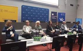 Pokaż zdjęcie: Prelegenci uczestniczący w panelu dyskusyjnym siedzą na czterech sofach ustawionych w rzędzie. Wśród panelistów jest pięć kobiet oraz dwóch mężczyzn. Jedna z kobiet, która nosi okulary, przemawia do zgromadzonych osób, trzymając w ręce mikrofon.