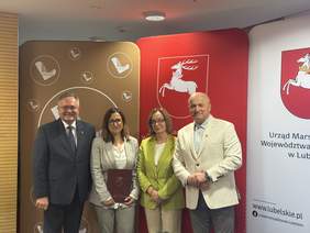 Pokaż zdjęcie: Na zdjęciu cztery osoby pozują do wspólnego zdjęcia podczas oficjalnego wydarzenia. Stoją przed ścinką z herbami i logotypami województwa lubelskiego. Jedna z kobiet trzyma bordową teczkę z dokumentem. W tle widoczne są symbole województw lubelskiego oraz napis "Urząd Marszałkowski Województwa Lubelskiego"