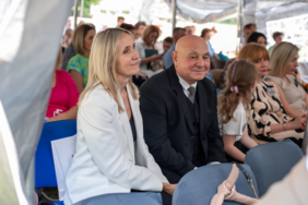 Pokaż zdjęcie: Zdjęcie zaproszonych gości siedzących na krzesełkach, zbliżenie na parę, mężczyznę w garniturze oraz młodą kobietę ubraną w białą marynarkę.