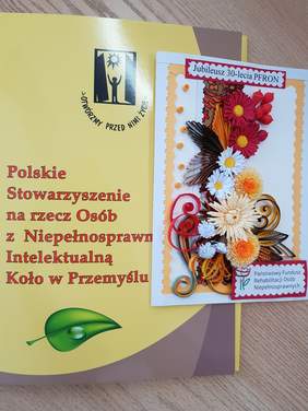 Na zółtym tle czerwony napis pełna nazwa PSONI koło w Przmyslu wraz z logiem w kolorze czarnym - postać unosząca ręce w otwartych okiennicach nad nią słońce. Z prawej strony przednia strona laurki przyozdobiona kolorowymi kwiatami