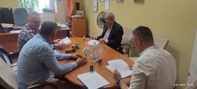 Pokaż zdjęcie: Przy owalnym stole siedzi czterech mężczyzn. Trzech panów trzyma w dłoniach dokumenty. Na stole leżą dokumenty, pieczątki, długopisy, telefony, woda mineralna. 