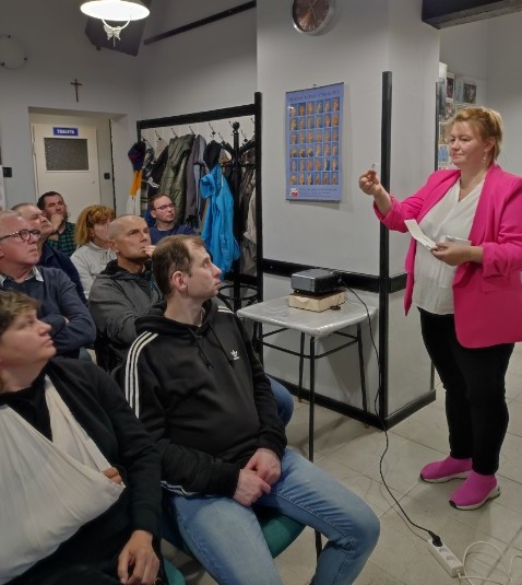 Pokaż zdjęcie: 1.	Grupa osób siedzi w sali wykładowej i patrzy na kobietę opowiadającej o aparacie słuchowym, który trzyma i pokazuje w prawej dłoni. W sali widać stojak na kurtki, mały stolik, na ścianie zegar i tablica z tłumaczeniem alfabetu na język migowy.