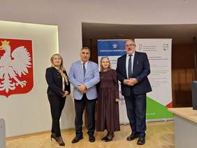 Pokaż zdjęcie: Cztery osoby stoją razem na tle godła Polski oraz banerów Państwowej Inspekcji Pracy i Samorządu Województwa Pomorskiego. Wszyscy są elegancko ubrani i pozują do wspólnego zdjęcia w sali konferencyjnej.