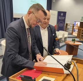 Pokaż zdjęcie: Dwóch mężczyzn w garniturach przy mównicy w auli Politechniki Koszalińskiej podpisuje porozumienie o współpracy. 