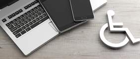 Pokaż zdjęcie: Na biurku leżą: laptop, tablet i telefon komórkowy. Po prawej stronie znajduje się jeszcze symbol osoby jeżdżącej na wózku inwalidzkim.  