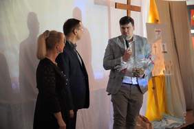 Pokaż zdjęcie: Na zdjęciu znajduje się dwóch mężczyzn oraz kobieta. Mężczyzna w młodym wieku trzyma w ręku mikrofon i przemawia