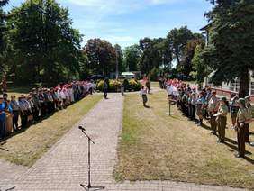 Pokaż zdjęcie: Na terenie zielonym stoją harcerze. Ustawieni są w dwóch długich rzędach. Na środku stoi mikrofon. W tle widać budynki i drzewa. 