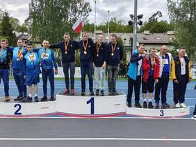 Pokaż zdjęcie: Na podium stoją trzy drużyny sportowe. Zawodnicy mają założone na szyjach medale. W tle widać flagę Polski. 