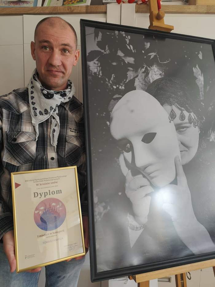 Pokaż zdjęcie: Osoba stojąca obok dużej, czarno-białej fotografii umieszczonej na sztaludze, trzymająca w dłoni dyplom za zdobycie pierwszego miejsca w konkursie plastycznym.