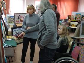 Pokaż zdjęcie: Na pierwszym planie mężczyzna w swetrze wskazuje palcem omawianą pracę. Na drugim planie widać dwie kobiety, jedna stoi i coś mówi. Druga, na wózku zastanawia się nad oceną. Wokół stoją różne prace artystyczne.