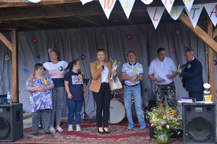 Pokaż zdjęcie: Na scenie stoi siedem osób, pośrodku nich kobieta z mikrofonem, która przemawia 