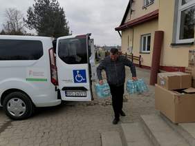 mężczyzna niesie butelki z wodą, z lewej strony stoi biały bus, z prawej widać front budynku WTZ