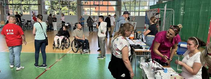 Pokaż zdjęcie: Na Sali gimnastycznej stoją stanowiska przy których siedzą przedstawiciele instytucji przed nimi stoją ludzie, dwie osoby poruszają się na wózkach inwalidzkich. Po prawej stronie kobieta sprawdza wyniki badań i rozmawia z osobą zainteresowaną. 