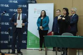Pokaż zdjęcie: Zdjęcie podczas briefingu prasowego, od lewej mężczyzna w garniturze, koło niego stoi kobieta z ciemnymi długimi włosami w niebieskiej marynarce, mężczyzna w garniturze, kobieta tłumacz języka migowego oraz mężczyzna w okularach z siwymi włosami ubrany w elegancki garnitur 