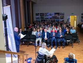 Widok ze sceny za grupę zebranych osób, które siedzą na krzesłach ustawionych w rzędach, z lewej strony mężczyzna przemawia, z przodu widać dwie osoby na wózkach inwalidzkich