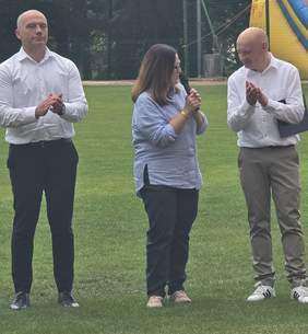Pokaż zdjęcie: Osoba przemawia do mikrofonu na stadionie podczas turnieju bocci, obok stoją uczestnicy i klaszczą. W tle widoczna dmuchana atrakcja oraz ogrodzenie boiska i drzewa.
