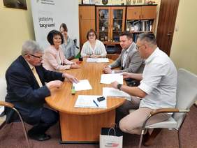 Pokaż zdjęcie: W gabinecie dyrektora znajduje sie stół, przy którym siedzi pięć osób: dwie kobiety i trzech mężzyzn. Podpisują umowę. Na stole leżą różne dokumenty, pieczątki i długopisy. 