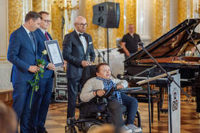 Pokaż zdjęcie: Na zdjęciu ceremonia wręczenia nagrody, która odbywa się w eleganckim, bogato zdobionym wnętrzu z marmurowymi kolumnami i złoceniami. Na pierwszym planie widzimy kobietę na wózku inwalidzkim, przemawiającą do mikrofonu. Obok niej stoi trzech mężczyzn w garniturach – jeden trzyma certyfikat, drugi różę a trzeci statuetkę. W tle widać fortepian. 