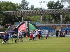 Pokaż zdjęcie: 13 zawodników trzyma w rękach łuki i mierzą  do tarczy. W tle  stadion.