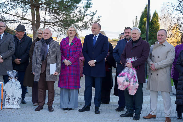 Grupa osób stoi na zewnątrz na wybrukowanym placu. Na pierwszym planie stoją w rzędzie trzy kobiety i sześciu mężczyzn, ubranych w eleganckie, jesienno-zimowe płaszcze. Na skraju po prawej mężczyzna trzyma przezroczystą torbę z różowymi kwiatami. Na ziemi po lewej widać dużą, owiniętą, podarunkową paczkę.