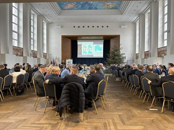 Pokaż zdjęcie: Duża sala, z narysowanym niebem na suficie. W trzech rzędach ustawione długie stoły, przy których siedzą elegancko ubrane osoby. Przed nimi stoi mężczyzna, który mówi do mikrofonu i wskazują na wiszący ekran.