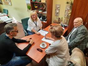 Pokaż zdjęcie: Przy owalnym stole siedzą dwie kobiety i dwóch mężczyzn. Rozmawiają ze sobą i uśmiechają się do siebie. Na stole leżą różne dokumenty, pieczątki, długopisy, telefony.  