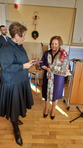 Na zdjęciu stoją dwie kobiety - jedna, po lewej trzyma w ręku medal, który zamierza wręczyc drugiej kobiecie - Dyrektor Kujawsko-Pomorskiego Oddziału PFRON Aleksandrze Gierej. Pani Dyrektor trzyma w rękach bukiet kolorowych kwiatów.