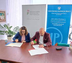 Pokaż zdjęcie: Przy stole konferencyjnym dwie osoby kobieta i mężczyzna podpisują umowy. W tle stoją rollupy z logo PFRON i Inspekcji Pracy.