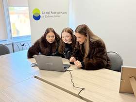 Trzy młode dziewczyny ubrane w futra i kurtki patrzą skupione w ekran laptopa, a za nimi logo Urzędu Statystycznego w Zielonej Górze