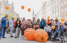 Pokaż zdjęcie: Tłum ludzi idzie ulicą Gdańska w Paradzie. Uczestnicy niosą jasnopomarańczowe parasole i balony. W centrum widać uśmiechnięte kobiety w marynarkach i kolorowych szalach. Grupa osób pozuje na Długim Targu w Gdańsku podczas Parady. Uczestnicy trzymają pomarańczowe balony i parasole. W tle widać zabytkowe kamienice i wieżę Ratusza. Wśród zgromadzonych są osoby na wózkach i uśmiechnięci opiekunowie.