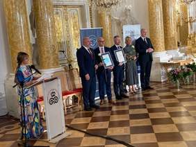 Pokaż zdjęcie: Czterech panów w ciemnych garniturze oraz pani w szarej garsonce. Z boku prowadząca galę w kolorowej sukni  Beata Chmielowska-Olech