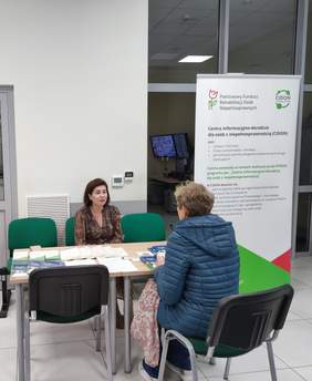 Pokaż zdjęcie: Kobieta zasiada przy stole naprzeciwko przedstawicielki PFRON, która udziela informacji i porad. Na stole leżą ulotki i materiały informacyjne. W tle banery informacyjne PFRON (CIDON).