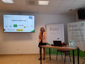 Pokaż zdjęcie: Sala konferencyjna, zdjęcie podczas konferencji, kobieta blondynka w czarnej sukience i łososiowej marynarce prowadzi wykład. Na ścianie slajd z prezentacji.
