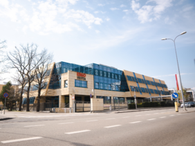 Pokaż zdjęcie: Budynek z przeszkloną elewacją i skośnymi panelami przeciwsłonecznymi. Nowoczesna architektura przy szerokiej ulicy, z logo firmy widocznym na froncie.