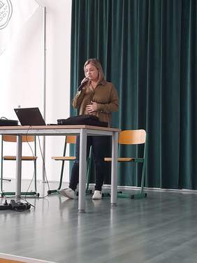Pokaż zdjęcie: Prelekcję wygłasza kobieta w brązowej bluzce i czarnych spodniach.