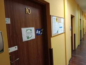 Pokaż zdjęcie: W korytarzu są drzwi, na których wisi tabliczka z informacją: CIDON w Oddziale PFRON. W tle widać drzwi do innych pokoi. Wszystkie są zamknięte. 