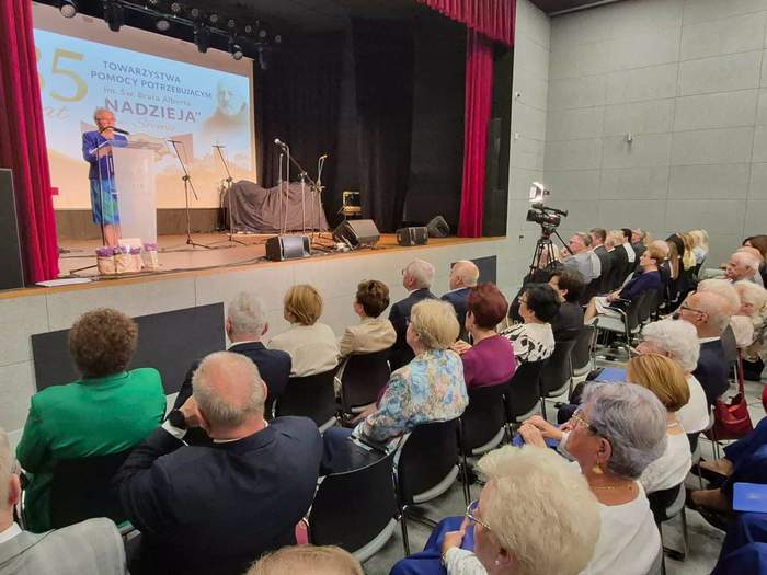 Pokaż zdjęcie: Widok na salę widowiskową, gdzie na scenie za jasną mównicą przemawia osoba w niebieskim stroju. Za nią, na dużym ekranie, wyświetla się napis 35 lat Towarzystwa Pomocy Potrzebującym im.. św. Alberta „NADZIEJA”. Scenę otaczają czerwone kurtyny, a obok stoi kamera na statywie. Na widowni w czarnych krzesłach siedzi elegancko ubrana, głównie starsza publiczność.