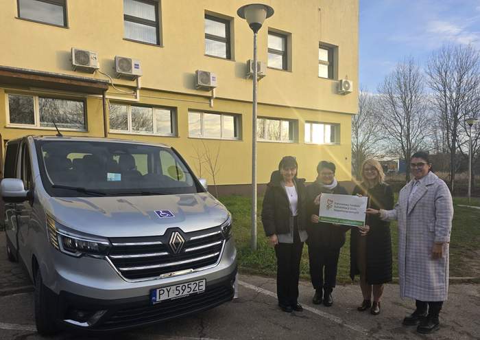 Pokaż zdjęcie: Cztery kobiety trzymają w dłoniach naklejkę z logo PFRON informującym o dofinansowaniu ze środków PFRON. Obok zaparkowany został srebrny bus. W tle kilkukondygnacyjny budynek oraz drzewa pozbawione liści na tle nieba z niewielką ilością chmur.