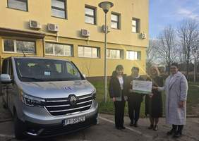 Pokaż zdjęcie: Cztery kobiety trzymają w dłoniach naklejkę z logo PFRON informującym o dofinansowaniu ze środków PFRON. Obok zaparkowany został srebrny bus. W tle kilkukondygnacyjny budynek oraz drzewa pozbawione liści na tle nieba z niewielką ilością chmur.