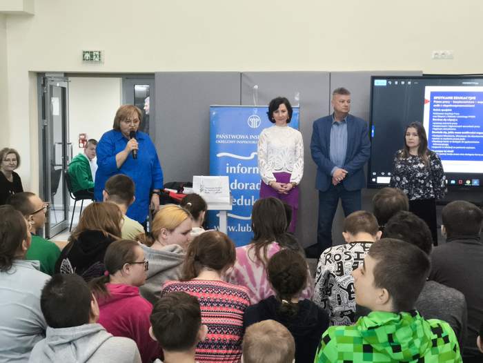 Pokaż zdjęcie: Sala gimnastyczna, młodzież siedzi na ławeczkach, przed nimi stoją trzy kobiety i jeden mężczyzna. Jedna z kobiet w niebieskiej bluzce trzyma w ręku mikrofon.