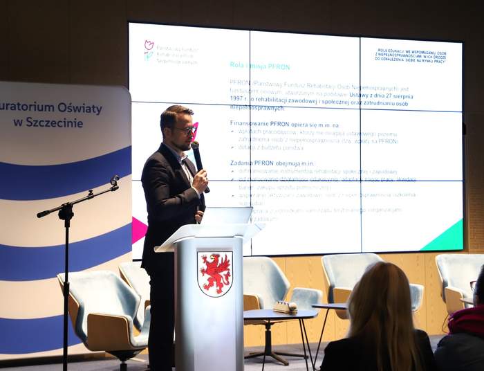 Pokaż zdjęcie: Zdjęcie przedstawia mężczyznę w garniturze stojącego za mównicą z godłem, prawdopodobnie podczas prezentacji. Na mównicy widnieje herb, przypominający herb Szczecina lub województwa zachodniopomorskiego (czerwony gryf). Za mężczyzną znajduje się duży ekran, na którym wyświetlana jest prezentacja w języku polskim, zawierająca nagłówki "Rola i misja PFRON" oraz informacje o roli, finansowaniu i zadaniach PFRON (Państwowy Fundusz Rehabilitacji Osób Niepełnosprawnych). Po lewej stronie mównicy stoi baner z napisem "Kuratorium Oświaty w Szczecinie" oraz flaga Unii Europejskiej. Na dole ekranu widoczne są fragmenty publiczności, w tym kobieta siedząca tyłem.