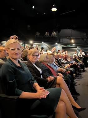 Pokaż zdjęcie: Dwie kobiety siedzą na widowni Białostockiego Teatru Lalek podczas XXVIII Przeglądu Twórczości Artystycznej Osób Niepełnosprawnych. W tle widać licznie zgromadzoną publiczność i uczestników wydarzenia.
