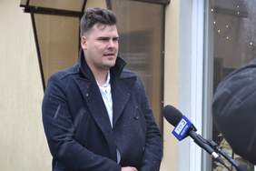 Pokaż zdjęcie: Wywiad prowadzony jest na dworze. Pada deszcz ze śniegiem. Dyrektor PFRON jest w ciemnym płaszczu, spod którego wystaje biała koszula. W jego stronę skierowany jest mikrofon ze znakiem TVP 3