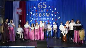 Pokaż zdjęcie: Na scenie występują dzieci z okazji 65-lecia Specjalnego Ośrodka Szkolno-Wychowawczego w Koluszkach. Dzieci śpiewają i grają na instrumentach. Mają na sobie eleganckie ubrania. 