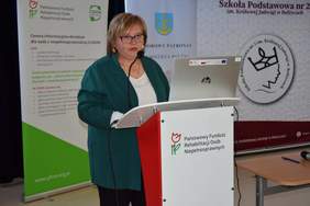 Pokaż zdjęcie: Przy mównicy stoi kobieta w zielonym swetrze. Na mównicy jest logo Funduszu.