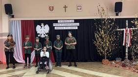 Pokaż zdjęcie: Grupa młodych osób i jedna osoba na wózku inwalidzkim wykonują pieśń patriotyczną na scenie, ubrani w stroje wojskowe z czerwonymi kwiatami w dłoniach. W tle biało-czerwona dekoracja z orłem i napis „Jestem Polakiem”, obok brzozowy krzyż z biało-czerwoną wstążką.