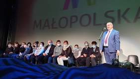 Pokaż zdjęcie: Scena "Opery Krakowskiej". Na środku stoi minister Paweł Wdówik, przemawia na scenie. Mężczyzna jest niewidomy, obok niego stoi pies przewodnik. W tle Laureaci konkursu plastycznego siedzą na krzesłach.
