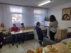 pod oknem za stolikiem siedzą 3 kobiety, na środku siedzi mężczyzna przed nim sprzęt elektroniczny, obok kobieta coś mu tłumaczy